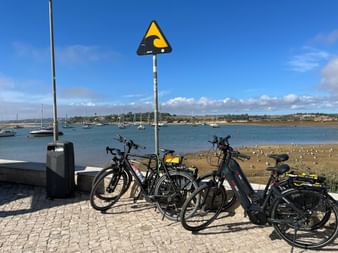 Drei Fahrräder auf einer Kopfsteinpflaster-Promenade in Alvor mit Hafenblick, Segelbooten und Sandstrand unter blauem Himmel.