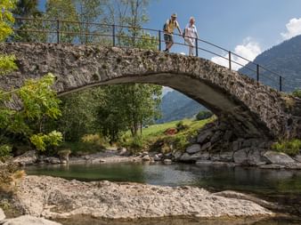 Zwei Wanderer stehen auf einen Bogenbrücke aus Steinen, mit einem Metallgeländer, die über einen schmalen Fluss führt.