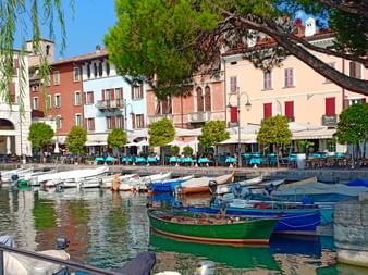 Petit port du lac de Garde avec des bateaux colorés amarrés dans une eau calme. Bâtiments méditerranéens aux volets rouges bordent le front de mer.