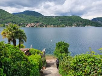 Steinstufen führen durch üppige Vegetation zum Lago Maggiore in Morcote, mit Palmen und Bergen rund um den See.