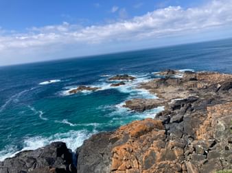 Zerklüftete Felsküste in Cornwall mit Wellen, die gegen dunkle und orangebraune Felsen schlagen. Türkisfarbenes Meer erstreckt sich zum Horizont.