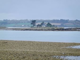 Kleine felsige Insel mit weißem Haus und Bäumen im ruhigen Wasser bei Tréguier, Bretagne. Kieselstrand im Vordergrund, ländliche Küste im Hintergrund.