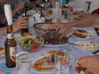 Tisch mit albanischen Gerichten: Grillfleisch, frischer Salat, Fladenbrot im Korb und Elbar-Bierflaschen. Mehrere Hände greifen nach Essen.