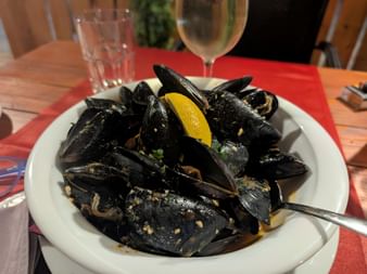 Weiße Schüssel mit schwarzen Muscheln und Zitronenschnitz, serviert mit Weißwein und Wasserglas auf roter Tischdecke an Bord der MS Dalmatino.