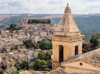 Vue panoramique de Ragusa Ibla avec clocher en pierre au premier plan et ville historique sur colline avec bâtiments traditionnels.