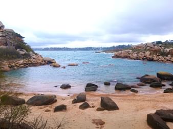 Sandstrand mit rosa Granitfelsen bei Ploumanac'h an der Rosa Granitküste der Bretagne. Ruhiges türkisfarbenes Wasser und felsige Küste unter bewölktem Himmel.