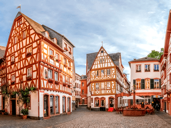 Kopfsteinpflasterplatz in Mainz, umgeben von bunten Fachwerkhäusern mit rot-weißen Fassaden. Außenbestuhlung auf dem Platz sichtbar.