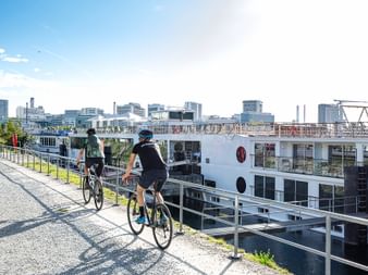 Des cyclistes roulent sur une route avec en arrière-plan un bateau sur le Rhin et la ville de Bâle.