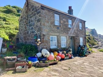 Traditionelles Steinhaus in Cornwall mit bunten Fischerbojen davor. Felsiger Hang hinter dem Haus unter blauem Himmel sichtbar.