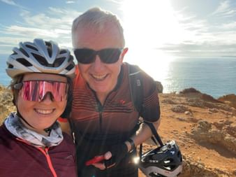 Zwei lächelnde Radfahrer in Radkleidung auf felsigen Küstenklippen nahe Ponte da Piedade, Algarve. Meer und blauer Himmel im Hintergrund.