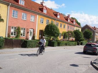Radfahrerin auf einer ruhigen Straße in Stockholm, gesäumt von bunten Reihenhäusern in Rosa und Gelb mit roten Ziegeldächern und grünen Hecken.