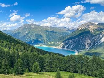 Lac alpin turquoise niché dans une vallée de montagne entourée de pentes boisées et de sommets spectaculaires sous un ciel bleu avec nuages blancs.