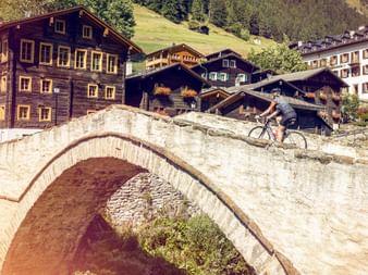 Une cycliste traverse un pont de pierre avec des maisons typiques du Valais en arrière-plan.