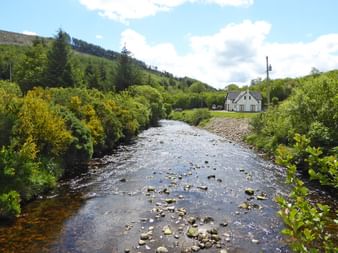 Weißes Landhaus neben einem steinigen Bach in den Wicklow Mountains, umgeben von üppiger grüner Vegetation und sanften Hügeln unter bewölktem Himmel.
