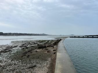 Geschwungener Meerwasserpool mit Betonmauer bei Ebbe bei Paimpol, Bretagne. Felsiger Strand und Küstenlinie unter bewölktem Himmel.