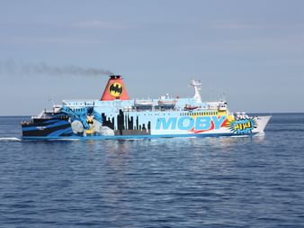 Weiße Moby-Fähre mit bunten Cartoon-Grafiken inklusive Batman-Logo am Schornstein, fährt auf ruhigem blauen Mittelmeer unter bewölktem Himmel.