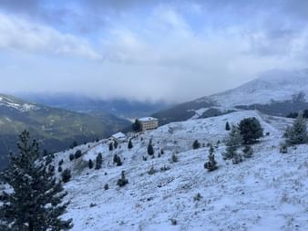 Hotel Weisshorn steht auf einem verschneiten Alpenhang mit vereinzelten Nadelbäumen. Berge und Täler unter bewölktem Himmel sichtbar.