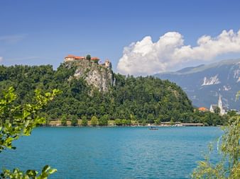 Burg Bled thront auf einem Felsen über dem türkisfarbenen Bleder See, mit Bergen im Hintergrund und einer Kirche am Ufer.