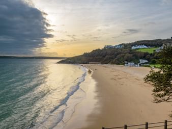 Plage de sable en Cornouailles au coucher du soleil avec eau turquoise, maisons blanches sur colline verte et ciel nuageux dramatique.