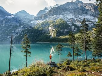 Randonneur en veste rouge debout sur un promontoire rocheux surplombant le lac turquoise d'Oeschinen entouré de sommets alpins enneigés.