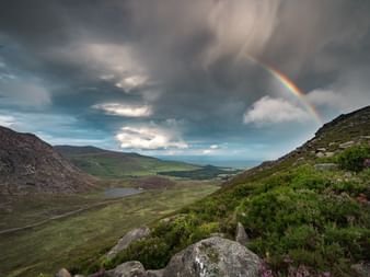 Regenbogen über grünen Hügeln und Tal in County Antrim, Nordirland. Dramatische Gewitterwolken über felsigen Hängen mit Heidekraut.