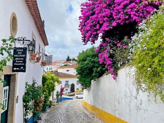 Schmale Kopfsteinpflasterstraße in Portugal mit weißen Gebäuden, leuchtend violetten Bougainvillea-Blüten über Mauern und Hotelschild.