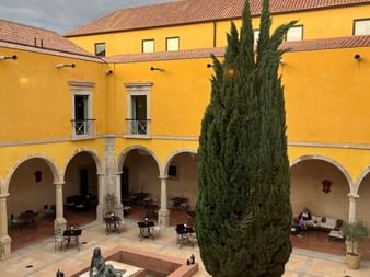 Innenhof der Pousada Convento Tavira mit gelben Wänden, Arkadengang, hoher Zypresse, Brunnen und Bronzeskulptur unter bewölktem Himmel.