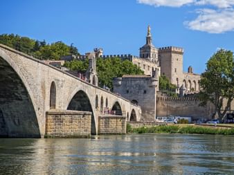 Pont historique d'Avignon en pierre avec plusieurs arches enjambant le Rhône, avec le Palais des Papes médiéval et les remparts derrière.