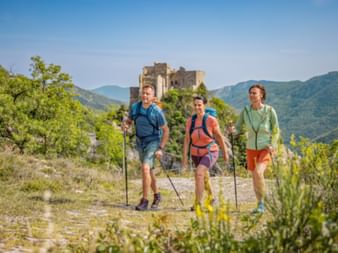 Trois randonneurs avec sacs à dos et bâtons sur un sentier de pierre avec le château de Castelvecchio sur une colline derrière eux.