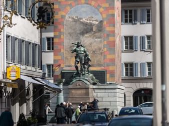 Bronze statue of William Tell with crossbow in Altdorf town square, surrounded by historic buildings with painted facade and pedestrians.