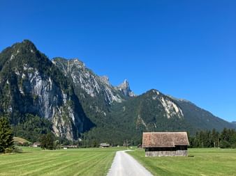 Gravel path through green meadows in Stockental valley near Bern, with traditional wooden barn and dramatic mountain peaks under clear blue sky.
