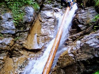 Drei Personen in Neoprenanzügen und Helmen seilen sich einen felsigen Wasserfall in einer engen Schlucht ab, umgeben von üppiger grüner Vegetation in Pfunds, Tirol.