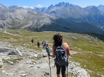 Wanderin mit Rucksack und Stöcken auf Bergpfad mit Blick auf Alpental. Gruppe voraus auf Weg mit dramatischen Gipfeln und türkisem See.