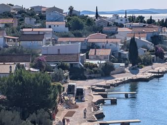 Mediterranes Küstendorf mit Häusern mit Terrakottadächern, Zypressen und einer Uferpromenade. Ein weißer Van ist am Ufer geparkt.