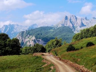 Schotterstraße schlängelt sich durch grüne Wiesen mit dramatischen felsigen Berggipfeln im Hintergrund unter teilweise bewölktem Himmel.