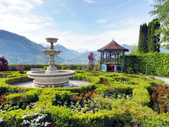 Verzierter Brunnen und Pavillon in gepflegtem Garten in Zell am See mit See und schneebedeckten Alpen im Hintergrund unter blauem Himmel.