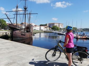 Radfahrerin mit Fahrrad am Hafen von Vila do Conde in Portugal, blickt auf historisches Holzschiff und Marina mit weißen Gebäuden im Hintergrund.