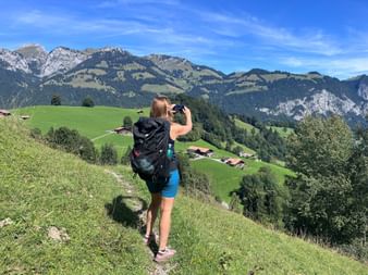Wanderin Sara mit Rucksack fotografiert Bergpanorama. Grüne Wiesen, verstreute Chalets und Alpengipfel unter blauem Himmel bei Grimmialp.