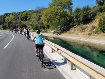 Gruppe von Radfahrern auf einer asphaltierten Straße entlang eines türkisfarbenen Kanals in Albanien. Bewaldete Hügel und blauer Himmel im Hintergrund.
