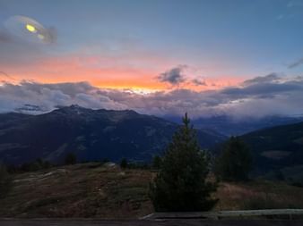 Dramatischer Sonnenuntergang mit orange-rosa Wolken über Alpengipfeln, vom Hotel Weisshorn aus mit Nadelbäumen im Vordergrund.