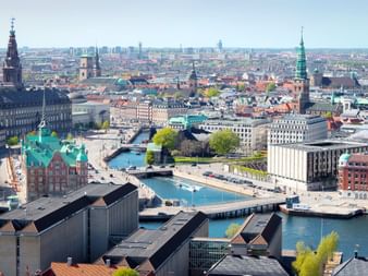 Luftaufnahme von Kopenhagen mit historischen Kirchtürmen, bunten Gebäuden, Wasserstraßen und Brücken unter blauem Himmel.