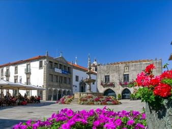 Historischer Stadtplatz in Viana do Castelo mit Steinbrunnen, bunten Blumen, mittelalterlichen Gebäuden und Straßencafé unter blauem Himmel.