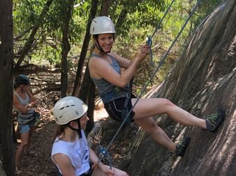Zwei Frauen mit weissen Helmen und Klettergurten erklimmen eine Felswand mit Seilen. Eine Frau klettert, die andere sichert unten zwischen Bäumen.