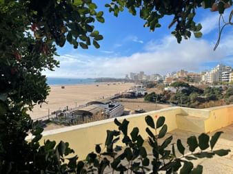 Blick auf den Strand Praia da Rocha in Portimão, umrahmt von grünem Laub, mit Sandstrand, Promenade und Küstengebäuden unter blauem Himmel.