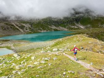Coureur de trail en maillot rouge court sur sentier vers lac alpin turquoise. Sommets brumeux et pentes vertes dans les Alpes suisses.