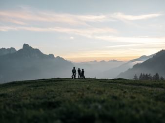 Silhouettes of four mountain bikers on a hilltop at sunset with layered mountain ranges and misty valleys in the background near Gstaad.