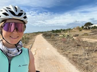 Lächelnde Radfahrerin Nina mit weißem Helm und lila Sonnenbrille auf einem sandigen Weg durch trockene Landschaft in der Algarve, Portugal.