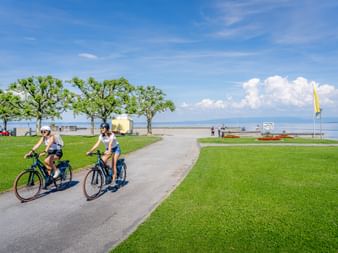 Deux cyclistes roulant sur un chemin pavé au bord du lac de Constance à Arbon. Pelouses vertes, arbres et ciel bleu avec nuages encadrent la scène.