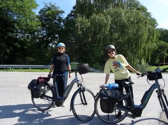 Zwei Radfahrer mit Helmen und Tourenrädern mit Packtaschen pausieren auf einer Straße bei Södertälje. Bäume und blauer Himmel im Hintergrund.
