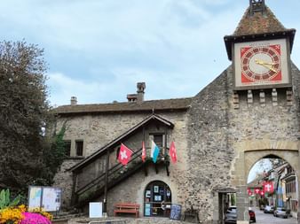 Mittelalterlicher Steinturm mit rotem Uhrenblatt in St-Prex. Schweizer Fahnen hängen am Gebäude, Kopfsteinplatz mit Brunnen und Blumen.